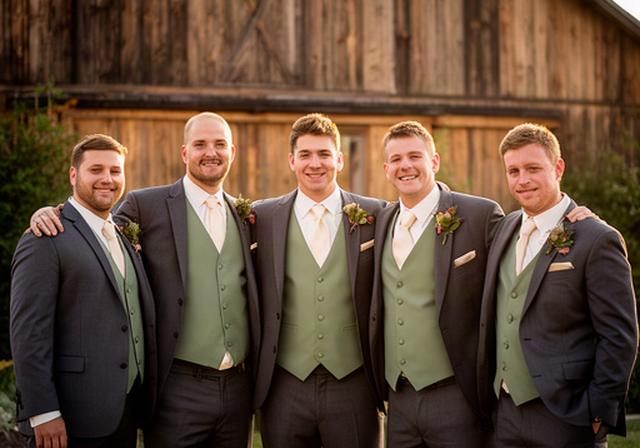 A wedding party of six men in coordinated three-piece suits with sage green vests posing outside a wooden barn venue
